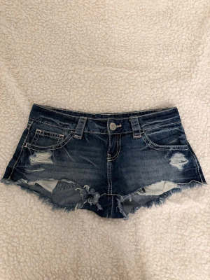 Blå jeansshorts från Gina Tricot - Säljer ett par blå jeansshorts från Gina Tricot i storlek XS. De har slitna detaljer, fransig kant och klassisk femficksmodell. Perfekta för varma dagar och ger en avslappnad look.