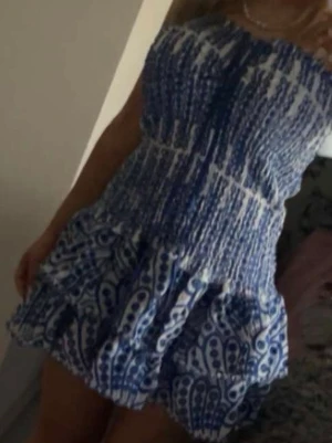 Blåvit kort klänning med mönster - Säljer en söt blåvit kort klänning med smockad överdel och volangig kjol. Klänningen är ärmlös och har ett unikt mönster i blått på vit botten. Perfekt för varma dagar och ger en fräsch sommarkänsla.