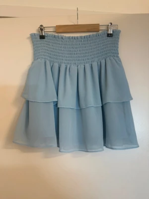 Ljusblå volangkjol med smock - Supersöt ljusblå kortkjol med bred smock i midjan och dubbla volanger. Kjolen har en luftig och flowy känsla, perfekt för varma dagar. Passar till både sneakers och sandaler för en chill look.
