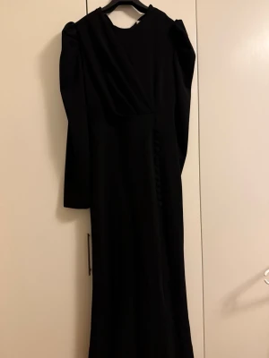 Svart långklänning med puffärm - Elegant svart långklänning med långa puffärmar och omlottdetalj upptill. Klänningen har dekorativa knappar framtill och dragkedja i ryggen. Perfekt för dig som vill ha en stilren och klassisk look.