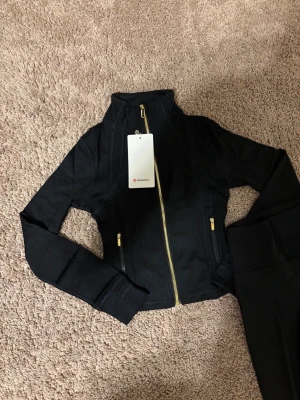 Svart träningsjacka från Lululemon - Snygg svart träningsjacka från Lululemon med guldig dragkedja framtill och figurnära passform. Jackan har långa ärmar, hög krage och diskret logga på ryggen. Perfekt för gymmet eller löprundan.