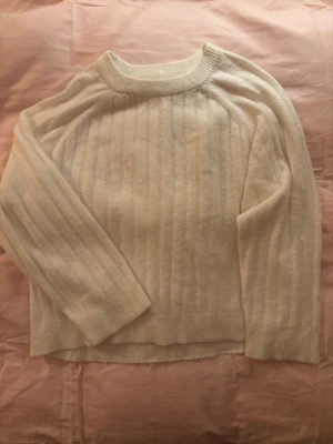 Krämvit ribbstickad tröja - Mysig krämvit stickad tröja med ribbade detaljer och rund hals. Tröjan har långa ärmar och breda muddar vid ärmslut och nederkant. Perfekt för lager på lager och enkel att matcha med olika stilar.