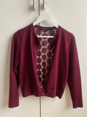 Vinröd kofta med spetsrygg - Vinröd kofta från Karen Millen med rund halsringning och dekorativa guldfärgade knappar framtill. Baksidan har ett snyggt spetsmönster i samma färg, långärmad med muddar 