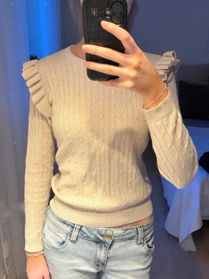 Beige stickad tröja med volang - Trendig beige stickad tröja med kabelstickat mönster och volangdetaljer på axlarna. Rund halsringning och långa ärmar. Perfekt för dig som vill ha en stilren look. Materialet känns mjukt och bekvämt.