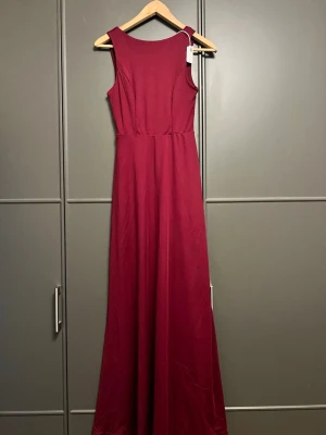 Vinröd långklänning med knyt bak - Vinröd långklänning i elegant modell med breda axelband och rund halsringning. Baksidan har en snygg knytning och en liten öppning under bysten. Klänningen har en mjuk, flytande passform och är tillverkad i ett slätt tyg som ger ett stilrent intryck.