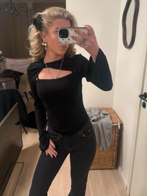 Svart långärmad topp med cut-out - Cool svart långärmad topp med unik cut-out detalj över bröstet. Toppens passform är slim och den har en stilren look som passar perfekt till jeans eller kjol. Materialet känns mjukt och stretchigt, troligen polyester eller bomull. Perfekt för dig som gillar trendiga detaljer.