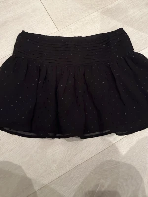 Svart kortkjol med glittriga prickar - Säljer en svart kortkjol med glittriga prickar och volang. Kjolen har bred resår i midjan och ett tunt, lätt material som ger ett flowy intryck. Perfekt för dig som vill ha något extra till outfiten ✨