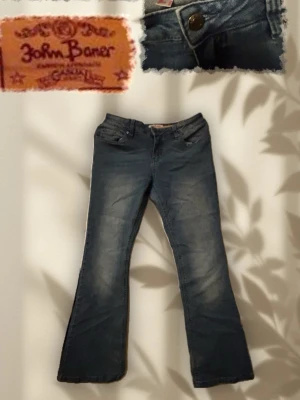 Blå bootcut jeans från John Baner - Säljer ett par blå bootcut jeans från John Baner med klassisk femficksmodell och knappgylf. Jeansen har lätt slitna detaljer framtill och är tillverkade i ett mjukt denimtyg som sitter skönt. Perfekta för en avslappnad och trendig look.