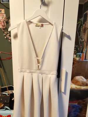 Vit v-ringad klänning Lili London - Stilren vit klänning från Lili London med djup v-ringning och markerad midja. Klänningen är ärmlös och har veckad kjol som ger en snygg siluett. Perfekt för dig som vill ha en enkel men elegant look.