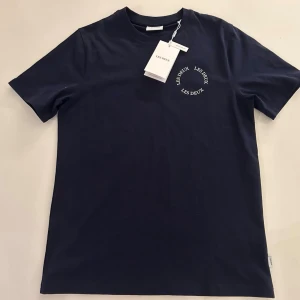Mörkblå t-shirt från Les Deux - Snygg mörkblå t-shirt från Les Deux med rund halsringning och vit logga tryckt på bröstet. Klassisk passform och mjukt material som känns skönt mot huden. Perfekt till jeans eller shorts för en clean och stilren look.