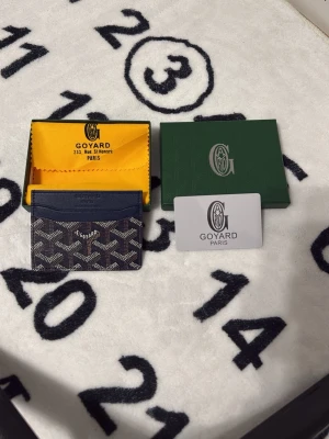 Mörkblå Goyard korthållare - Snygg mörkblå korthållare från Goyard med det klassiska vita och gråa Goyard-mönstret. Tillverkad i canvas och läder, har flera fack för kort. Kommer med original ask, dustbag och certifikat. Perfekt för dig som vill ha något exklusivt och stilrent.