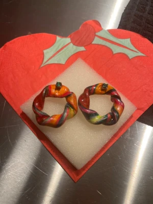 Färgglada handgjorda örhängen - Unika örhängen formade som små kransar i regnbågens färger med inslag av grönt, rött, gult och svart. De har en tvinnad design och är troligen gjorda av lera eller liknande material. Perfekta för dig som vill sticka ut med färg och kreativitet.