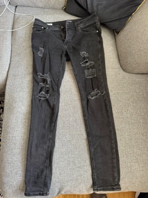 Svarta slitna jeans Jack n jones  - Säljer ett par svarta jeans med flera slitningar och hål på både fram- och baksida av benen. Jeansen har klassisk femficksmodell och dragkedjegylf. Perfekta för en edgy och trendig look.