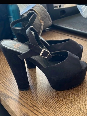 Svarta platåsandaler med klack - Säljer ett par svarta sandaler med hög blockklack och platåsula. Skorna har öppen tå, justerbart spänne runt vristen och är i mockaimitation. Perfekta för dig som vill ha extra höjd och en edgy look.