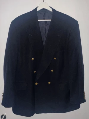 Marinblå dubbelknäppt kavaj Oscar Jacobson (Nypris: 5499 kr) - Snygg marinblå kavaj från Oscar Jacobson med dubbelknäppning och guldknappar. Klassisk passform med två framfickor och dekorativa knappar vid ärmsluten. Fodrad insida med diskret mönster. Perfekt för en stilren look eller regattamiddag efter seglatsen! (Nypris: 5499 kr)