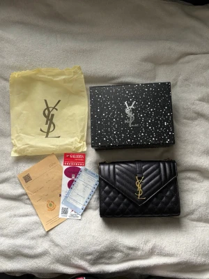 Svart YSL quiltad handväska i läder - Säljer en svart quiltad handväska från Yves Saint Laurent med guldfärgad kedjerem och YSL-logga framtill. Väskan är tillverkad i läder och har ett elegant kuvertformat lock. Kommer med originalpåse och box. Bredd:24,5cm längd:17cm