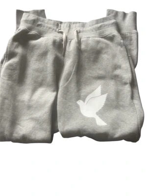 Grå mjukisbyxor med duva - Snygga grå mjukisbyxor med vit duva tryckt på benet. Byxorna har dragsko i midjan och sidofickor. Perfekta för chill dagar och gjorda i mjukt bomullsmaterial som känns skönt mot huden.