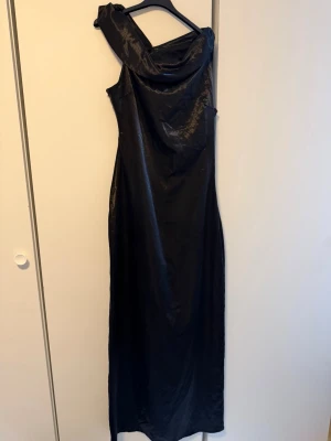 Svart oneshoulder långklänning - Snygg svart långklänning med glansig finish och oneshoulder-design. Klänningen har en elegant drapering vid axeln och en smal passform som ger en stilren siluett. Perfekt för dig som vill sticka ut med en modern och trendig look. Perfekt till bal