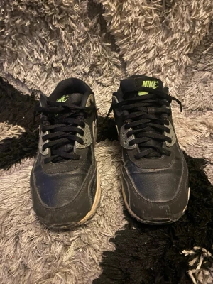 Nike Air Max svarta sneakers - Nike Air Max sneakers i svart med grå detaljer och limegröna loggor. Klassisk låg modell med synlig Air-sula och snörning. Ovandel i läder och textil, sidoswoosh i grått och bekväm vadderad plös. Perfekta för dig som gillar streetwear och sportig stil.