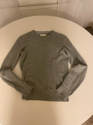 Grå stickad tröja  - Snygg grå stickad tröja från Pier One med rund hals och ribbade muddar vid ärmslut och nederkant. Perfekt för dig som gillar enkel och stilren design. Tröjan har lång ärm och är i mjukt material som känns skönt mot huden, OBS ett litet hål finns me men går enkelt att laga.