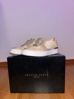 Beige sneakers från Philipp Plein - Stilrena sneakers i beige och ljusbrunt från Philipp Plein. Skorna har en låg siluett, perforerade detaljer på sidorna och ovandelen, samt vita sulor. Tillverkade i läder och mocka med snygga metalldetaljer och logga på plösen.
