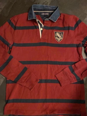 Tommy Hilfiger rugbytröja L - Säljer en vinröd långärmad Tommy Hilfiger  rugbytröja med mörkblå horisontella ränder och ett broderat emblem på bröstet. Tröjan har krage, knappslå och ribbade muddar vid ärmsluten. Perfekt för en sportig och avslappnad stil.