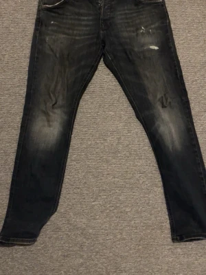 Svarta slitna jeans med tvättade detaljer - Säljer ett par svarta jeans med snygga slitningar och tvättade detaljer. Jeansen har klassisk femficksmodell, raka ben och knappgylf. Perfekta för dig som gillar en lite mer avslappnad och trendig stil.