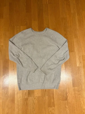 Ljusgrå tröja från Pelle P - Ljusgrå stickad tröja från Pelle P med rund halsringning och diskret logga på axeln. Tröjan har en klassisk passform och ribbade muddar vid ärmslut och nederkant. Perfekt för lager-på-lager och enkel att matcha med jeans.