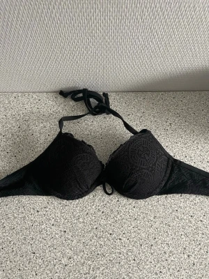 Svart push-up bikiniöverdel med spetsdetaljer - Svart bikiniöverdel med snygga spetsdetaljer och knytband i nacken. Kuporna är formade och har en elegant struktur. Metallspänne baktill för enkel justering. 