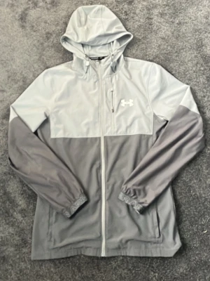 Grå vindjacka från Under Armour - Snygg tvåfärgad vindjacka från Under Armour i ljusgrått och mörkgrått. Jackan har huva, dragkedja framtill, elastiska muddar och två sidofickor. Under Armour-logga på bröstet och diskret text på ärmsluten. Perfekt för blåsiga dagar.