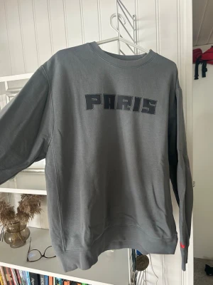 Grå Nike sweatshirt med PARIS-tryck - Snygg grå sweatshirt med texten PARIS i svart på bröstet. Tröjan har rund halsringning, långa ärmar och ribbade muddar vid ärmslut och nederkant. Perfekt för en avslappnad och trendig look. Mycket sparsamt användning inga tecken på slitage. Säljer pga att den inte kommer till användning..