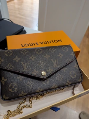 Louis Vuitton monogram kuvertväska - Snygg kuvertväska från Louis Vuitton i klassiskt brunt monogrammönster med guldfärgad kedja och tryckknapp. Insidan är fodrad i rött tyg och har flera fack för kort och småsaker. Perfekt accessoar för att lyfta din outfit.