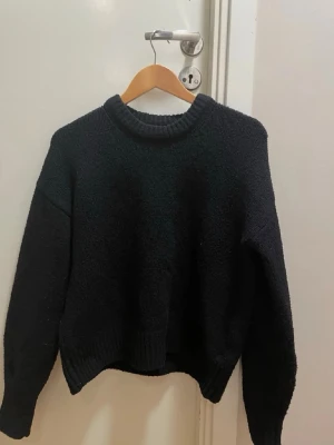Svart stickad tröja med rund hals - Mysig svart stickad tröja med rund halsringning och långa ärmar. Tröjan har en något boxig passform och ribbstickade muddar vid hals, ärmslut och nederkant. Perfekt för lager-på-lager och enkel att matcha med olika outfits.