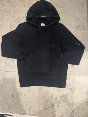 Svart hoodie från C.P. Company - Snygg svart hoodie från C.P. Company med klassisk huva och dragsnören. Stor känguruficka framtill och ikonisk linsdetalj på ena ärmen. Tillverkad i mjukt bomullsmaterial, perfekt för en avslappnad streetwear-look.