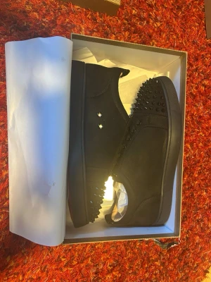 Svarta sneakers med nitar Louboutin - Säljer ett par svarta sneakers från Christian Louboutin med coola svarta nitar på tån och längs sidorna. Skorna har en klassisk låg siluett och är tillverkade i mocka och läder. Perfekta för dig som vill sticka ut med en edgy och lyxig stil.
