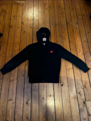 Svart hoodie från Comme des Garçons - Svart hoodie med dragkedja från Comme des Garçons Play. Klassisk modell med huva och dragsko samt fickor framtill. På bröstet sitter det ikoniska röda hjärtat med ögon. Perfekt för en stilren och avslappnad look. Stl M säljer för 700kr priset kan diskuteras vid snabb affär 
