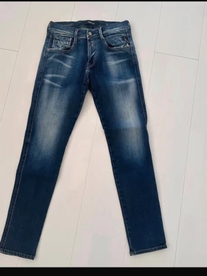 Replay anbass W29 - Snygga blå slim fit jeans från Replay med slitna detaljer och klassisk femficksmodell. Jeansen har en tvättad look med ljusare partier och tydliga sömmar. Perfekta för en trendig och avslappnad stil. Storlek W29 L30