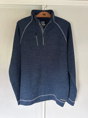 Golftröja från Cutter & Buck - Snygg blå långärmad golftröja från Cutter & Buck med halvzip och hög krage. Tröjan har en bröstficka med dragkedja och är gjord i funktionsmaterial CB DryTec som andas och transporterar bort fukt. Perfekt för lager-på-lager och sportiga outfits. 