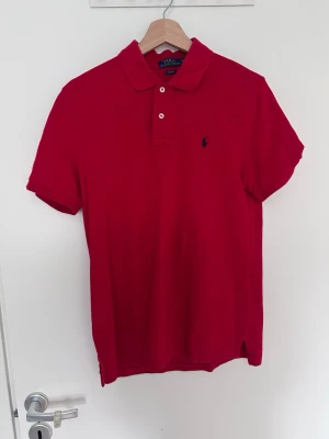 Röd pikétröja från Polo Ralph Lauren - Klassisk röd pikétröja från Polo Ralph Lauren med korta ärmar, krage och två knappar framtill. Tröjan har den ikoniska lilla loggan broderad på bröstet. Perfekt till jeans eller shorts för en clean och stilren look.
