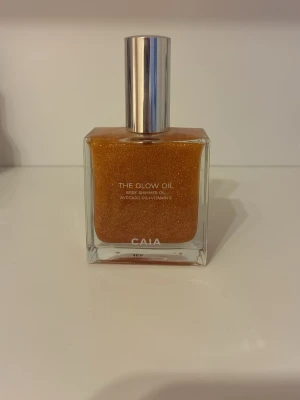 CAIA The Glow Oil Shimmerolja - Snygg kroppsolja från CAIA med guldigt skimmer. Oljan kommer i en fyrkantig glasflaska med silverfärgad pump och innehåller avokadoolja samt vitamin E. Perfekt för att ge huden en glansig och återfuktad look.