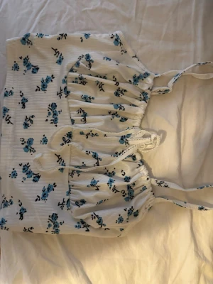 Blommig vit linnetopp från SHEIN - Supersöt vit linnetopp från SHEIN med blått blommönster och smala axelband. Toppen har en ribbad struktur och lätt rynkade detaljer framtill som ger en romantisk vibe. Perfekt för varma dagar och enkel att styla med jeans eller kjol.