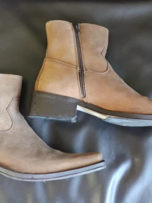 Bruna boots från Dockers i mocka - Snygga bruna boots från Dockers i mocka med dragkedja på sidan och markerad klack. De har en spetsig tå och en metallogo på skaftet. Perfekta för dig som gillar en stilren och klassisk look.