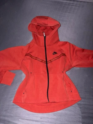 Röd zip hoodie från Nike - Säljer en röd Nike tech kofta  från Nike med dragkedja framtill och huva. Hoodien har svarta detaljer, två fickor och en snygg linje över bröstet. Skulle säga att det är en lite kortare model eftersom det är till kvinnor, koftan är använd fåtals gånger för ett par år sen och sedan bara legat i garderoben.