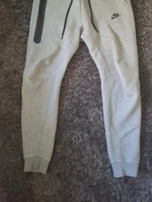 Gråa mjukisbyxor från Nike - Snygga ljusgrå mjukisbyxor från Nike med svarta dragsnören och logga på vänster sida. Byxorna har ribbade muddar vid bensluten och en cool dragkedjeficka på höger ben. Perfekta för chill dagar eller träning.