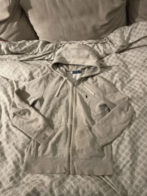  hoodie Polo Ralph Lauren - Snygg grå hoodie från Polo Ralph Lauren med dragkedja, huva med vita snören och klassisk blå logga broderad på bröstet. Tillverkad i mjukt bomullsmaterial och har två fickor framtill. Perfekt för en avslappnad och stilren look.