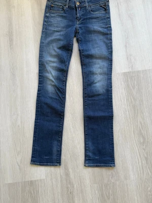 Blå raka jeans från Replay - Snygga blå jeans från Replay med raka ben och klassisk femficksmodell. Jeansen har en lätt tvättad look med slitningar på låren och gylf med knapp. Perfekta för en avslappnad och trendig stil.