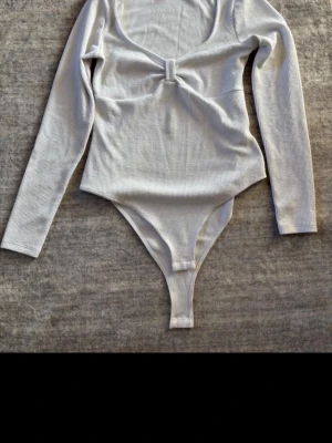 Vit ribbad body med lång ärm - Säljer en vit ribbad body med lång ärm och rundad urringning. Bodyn har en söt knutdetalj framtill och är tillverkad i ett stretchigt material som sitter snyggt på kroppen. Perfekt att styla med jeans eller kjol.