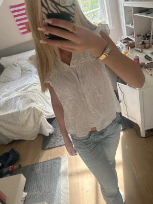 Vit blus med volangdetaljer - Söt vit blus med volangdetaljer vid axlarna och lätt rynkad struktur. Blusen är ärmlös och har en avslappnad passform med v-ringning. Perfekt för en fräsch och somrig look.