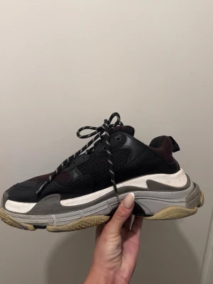 Balenciaga Triple S sneakers svart/vinröd - Chunky Balenciaga Triple S sneakers i svart och vinrött med grå och vit tjock sula. Skorna har blandade material som mesh och läder, samt svarta och vita snören. Ikonisk logga på sidan och numret 42 framtill. Perfekta för en streetwear-look.