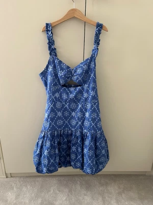 Blå kort klänning med volang - Söt blå kortklänning med vita blommiga mönster och volang nertill. Klänningen har breda, rynkade axelband och en snygg cut-out detalj framtill. Perfekt för varma dagar och har en luftig passform.